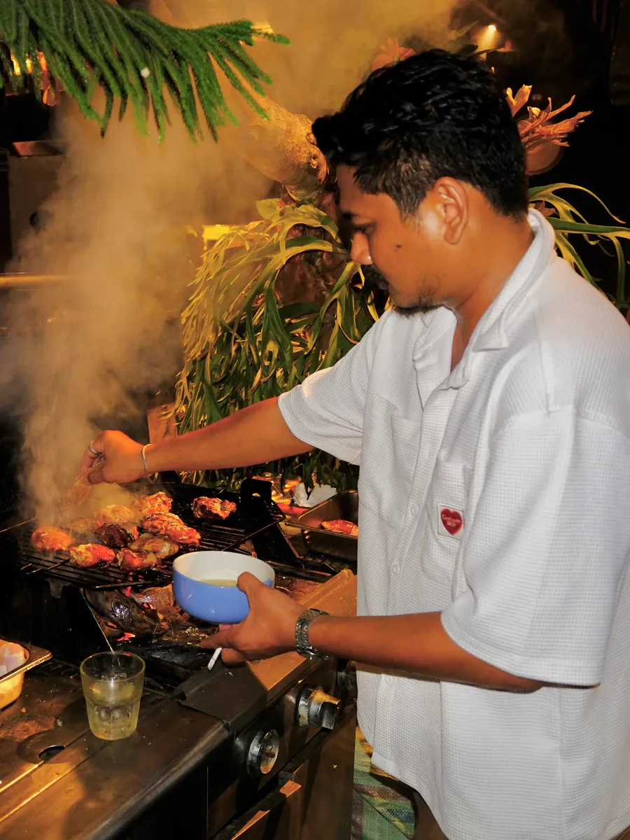 Staff grilling chicken on an outdoor barbecue at night, with smoke rising under tropical plants