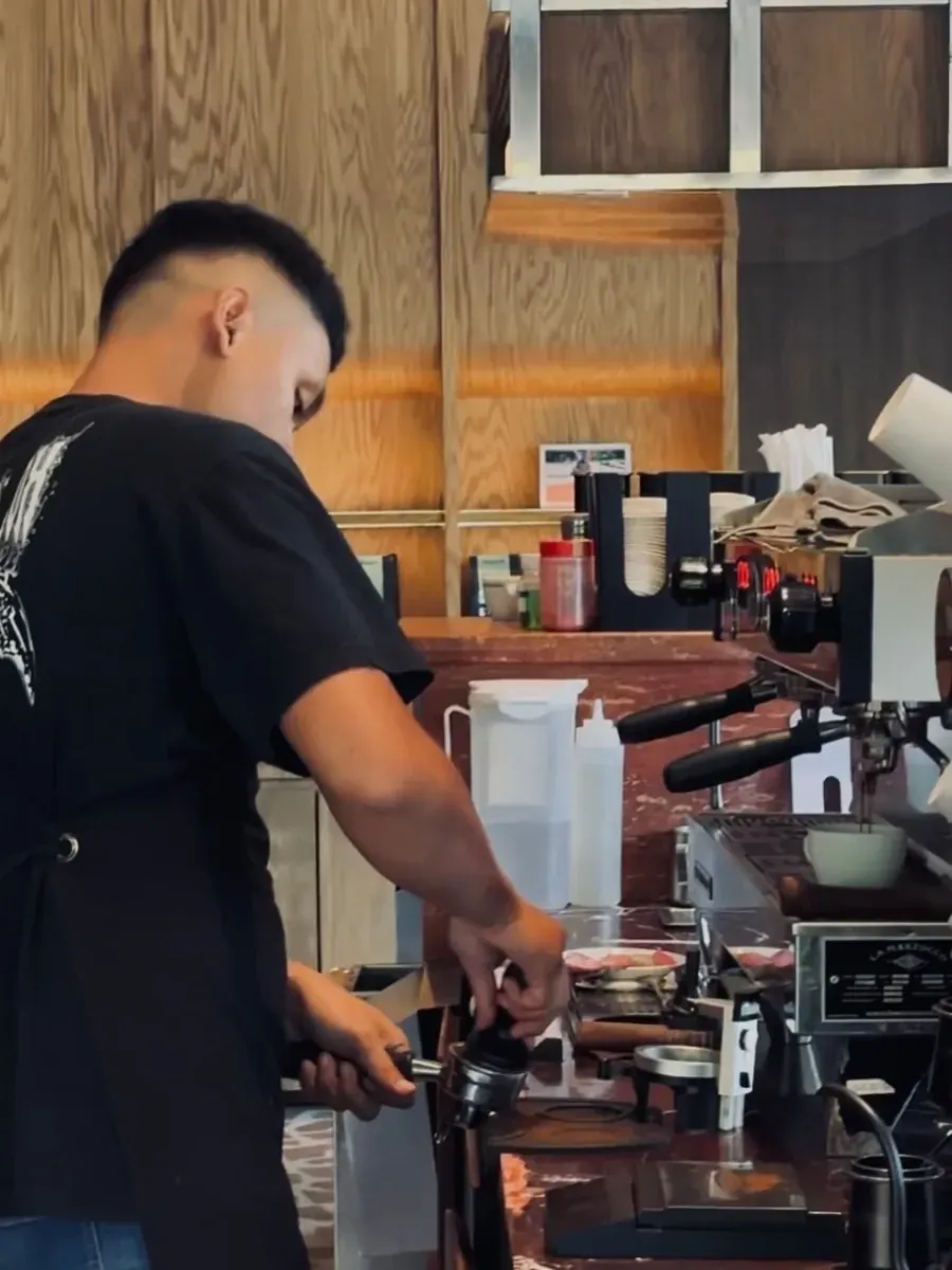 Barista tamping a portafilter beside a La Marzocco espresso machine pouring into a white cup