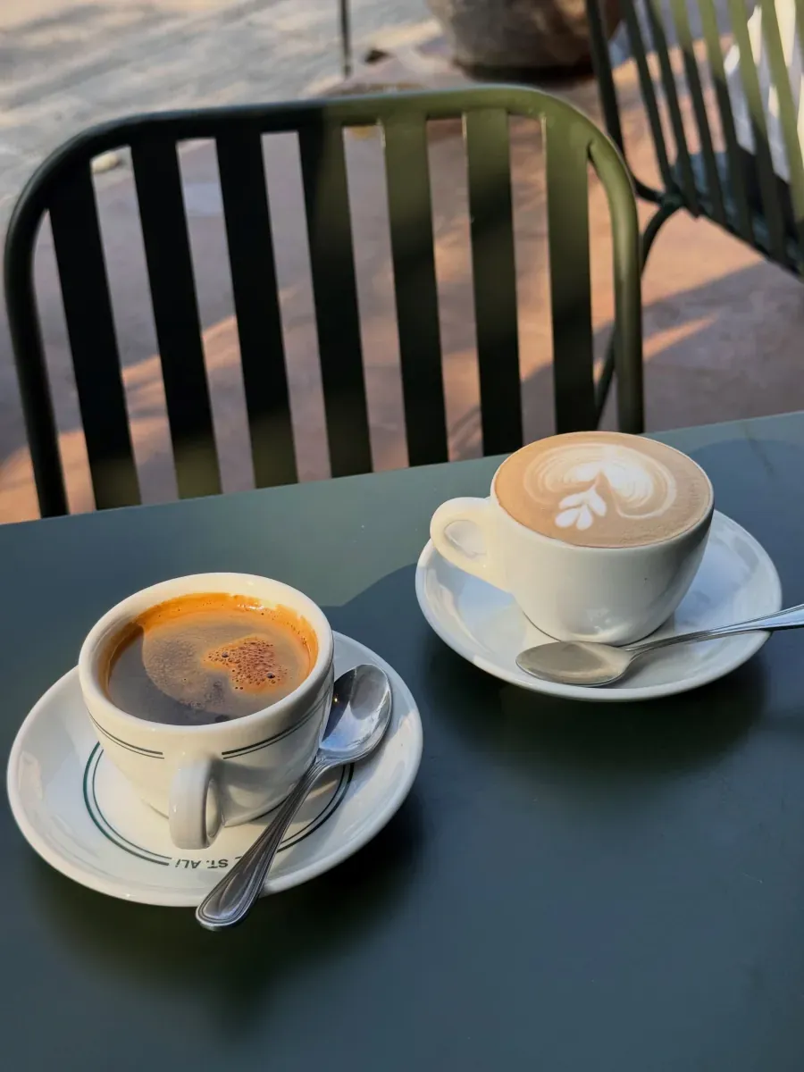 Black coffee in a green-banded ST. ALi cup beside a latte with heart art on a dark outdoor table in morning light