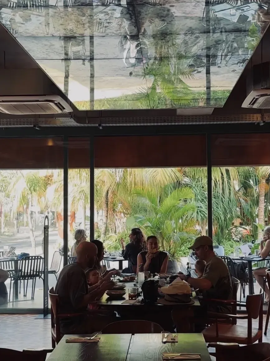 Guests at a long wooden table under the mirrored ceiling, with glass walls opening onto tropical greenery outside