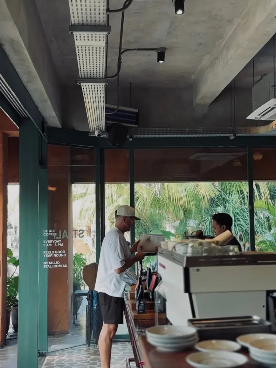 Customer at the wooden bar with the espresso machine and bean bags, ST. ALi hours on the glass, and palms outside