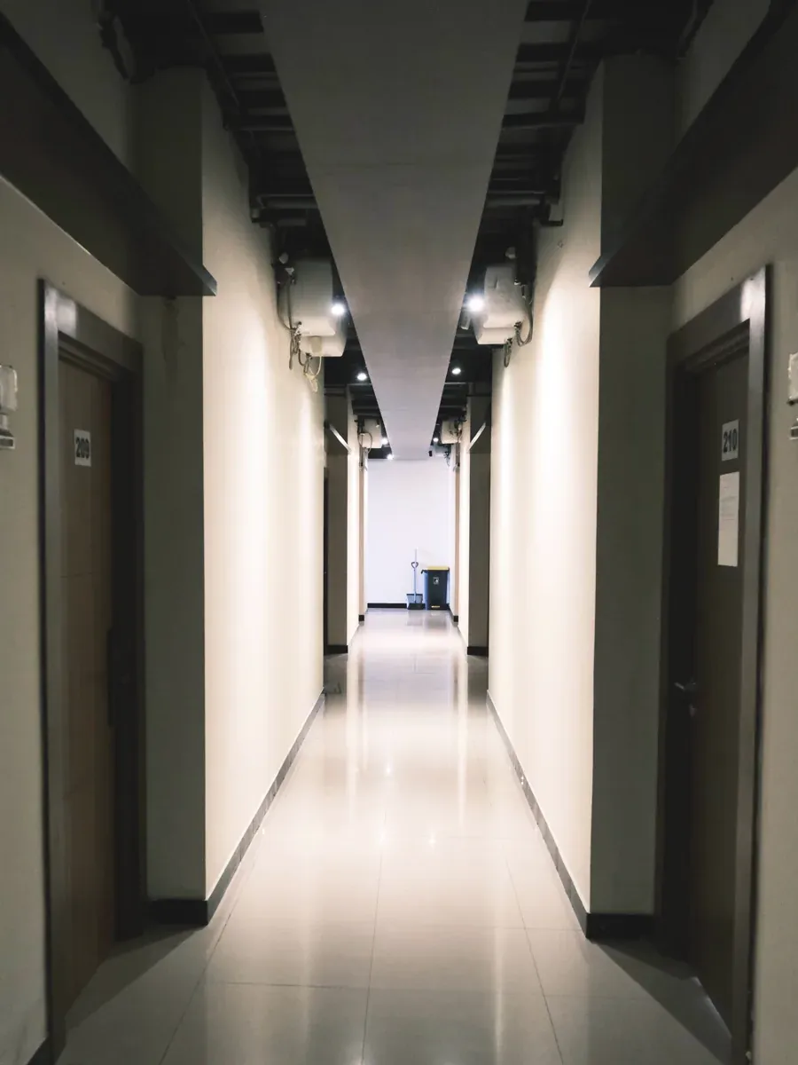 Long indoor corridor with wooden doors, ceiling duct, and spotlights on polished tile