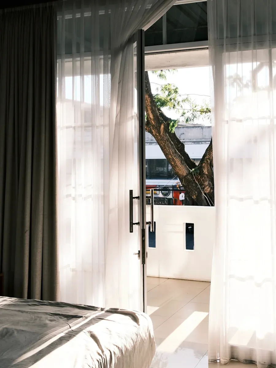 Sheer curtains and open glass doors looking out to a balcony tree and the street beyond
