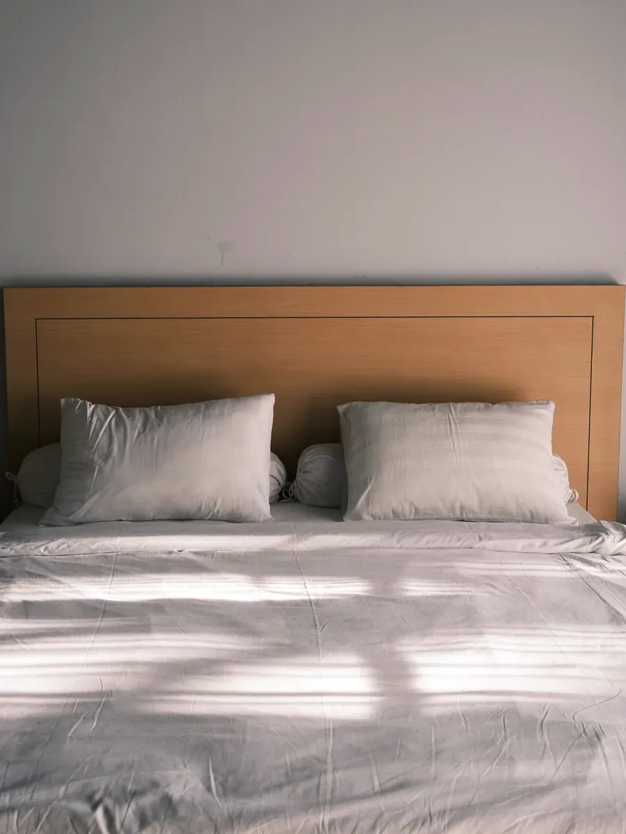 Neatly made bed with white pillows, bolsters, and a light wood headboard in morning light